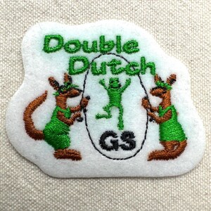 Puede incluir: Parche bordado con las palabras "Double Dutch" en hilo verde. El parche presenta dos figuras parecidas a canguros con trajes verdes sosteniendo cuerdas para saltar, y una rana verde saltando en el medio. Las letras "GS" est&aacute;n en un c&iacute;rculo.