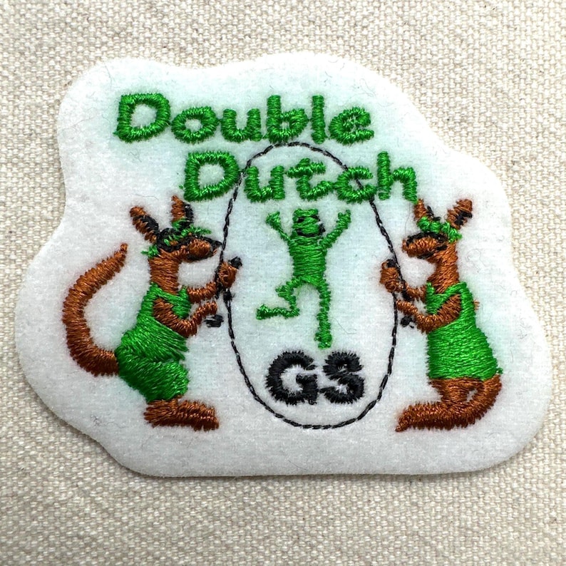 Puede incluir: Parche bordado con las palabras "Double Dutch" en hilo verde. El parche presenta dos figuras parecidas a canguros con trajes verdes sosteniendo cuerdas para saltar, y una rana verde saltando en el medio. Las letras "GS" est&aacute;n en un c&iacute;rculo.
