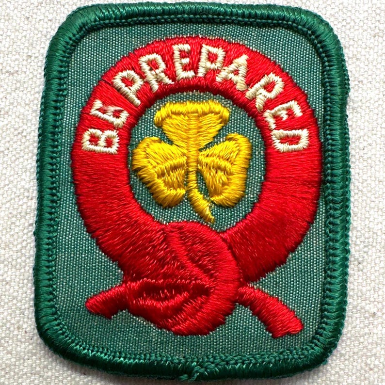 May include: Embroidered patch with a green border and background. The patch features a red circle with the words "BE PREPARED" in white. Inside the circle is a yellow trefoil. A red knot design is at the bottom.