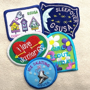 May include: A collection of embroidered Girl Scout patches. Designs include birdhouses, a heart with an arrow, a crescent moon, flowers, and an ice skating figure. Each patch has the text "GSUSA".