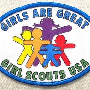 May include: Oval patch with a blue border featuring the text "GIRLS ARE GREAT" and "GIRL SCOUTS USA." The patch has a white background with colourful figures in yellow, orange, purple, red, and blue.