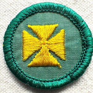 May include: Embroidered patch featuring a yellow cross design on a green background. The cross is centered within a circular patch with a green stitched border. The patch is likely for a uniform or craft project.