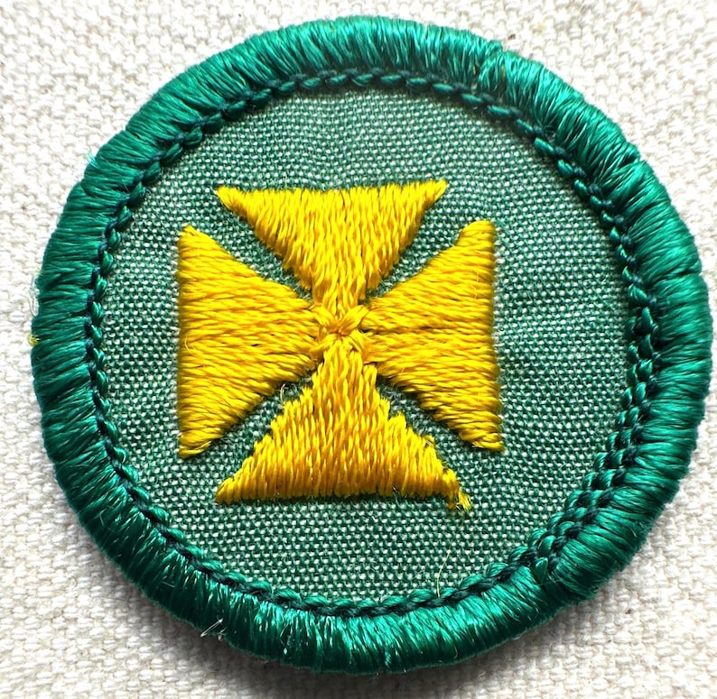 May include: Embroidered patch featuring a yellow cross design on a green background. The cross is centered within a circular patch with a green stitched border. The patch is likely for a uniform or craft project.