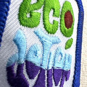 May include: Embroidered patch with the words "eco" and "delish" in green, blue, and purple thread. The "o" in "eco" is an avocado. The patch has a white background and a blue border.
