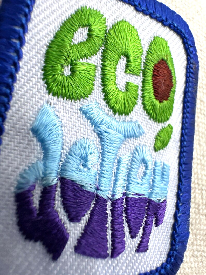 May include: Embroidered patch with the words "eco" and "delish" in green, blue, and purple thread. The "o" in "eco" is an avocado. The patch has a white background and a blue border.