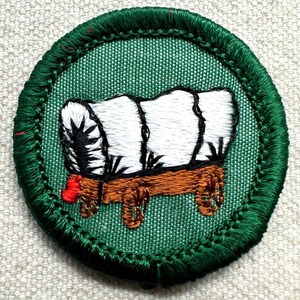 May include: Embroidered patch featuring a covered wagon design. The wagon has a white top, brown wooden base, and red accents. The patch is circular with a green border and green fabric background.
