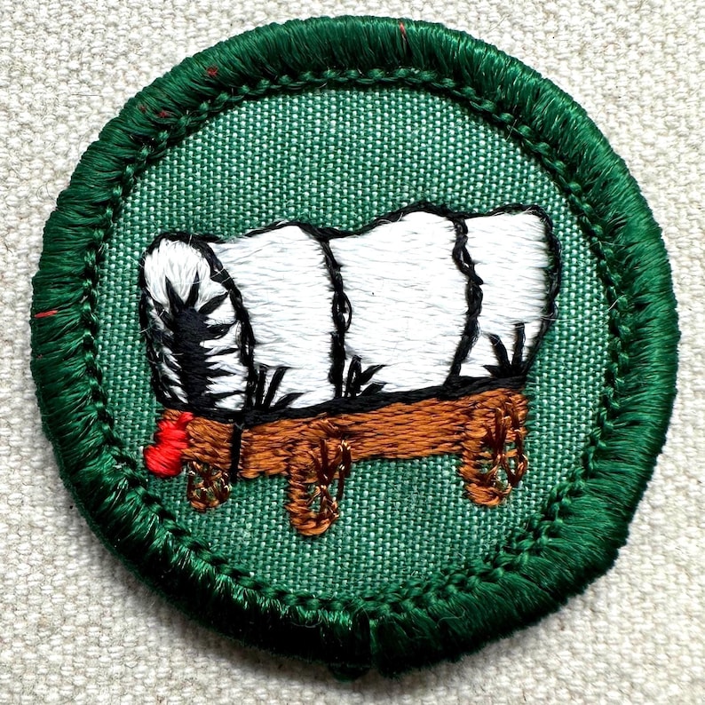 May include: Embroidered patch featuring a covered wagon design. The wagon has a white top, brown wooden base, and red accents. The patch is circular with a green border and green fabric background.