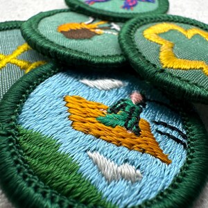 May include: A collection of embroidered patches with green borders. The patches feature various designs, including a person on a flying carpet, a yellow cross, and a yellow clover. The patches are on a white surface.