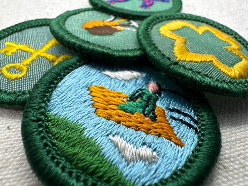 May include: A collection of embroidered patches with green borders. The patches feature various designs, including a person on a flying carpet, a yellow cross, and a yellow clover. The patches are on a white surface.