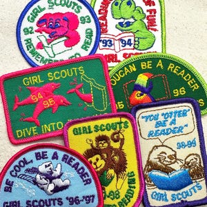 May include: A collection of Girl Scout patches with various designs and text. The patches feature images of animals, such as an elephant, dolphins, a toucan, a bear, and an otter, along with phrases like "Reading is Tons of Fun" and "Be Cool, Be a Reader."