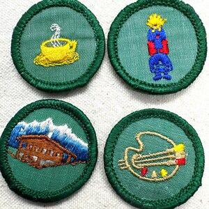 May include: Four circular embroidered patches with green fabric and dark green stitched borders. The designs feature a yellow teacup with steam, a blue and red figure with a yellow top, a brown cabin with blue mountains, and a gold artist palette.