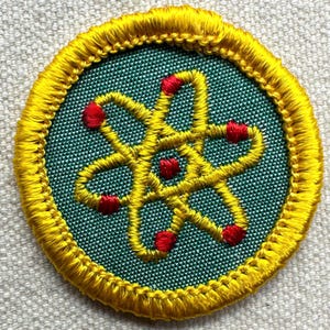 May include: Circular embroidered patch with a yellow atom design and red accents on a teal background. The patch has a yellow border. The design is machine-embroidered.