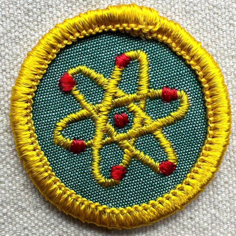May include: Circular embroidered patch with a yellow atom design and red accents on a teal background. The patch has a yellow border. The design is machine-embroidered.