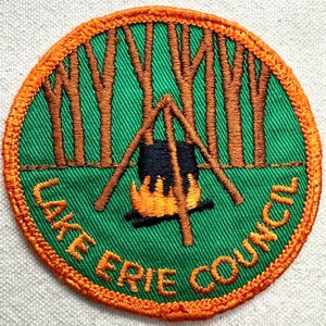 May include: A circular embroidered patch with an orange border on a green background. It features brown tree trunks, a black cooking pot suspended over a yellow and orange fire, and the text LAKE ERIE COUNCIL stitched in orange along the bottom.