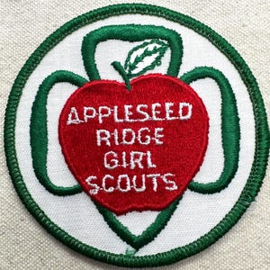 May include: A circular embroidered patch with a white background and a dark green stitched border. In the center, a red apple shape features the white embroidered text APPLESEED RIDGE GIRL SCOUTS, topped with a small green leaf icon.