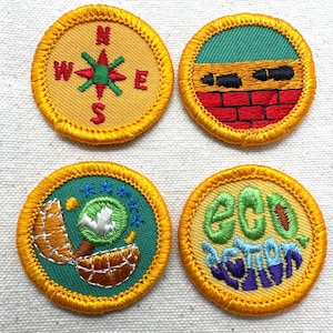 May include: Four embroidered patches with yellow borders. One features a compass rose, another a brick wall, a third a globe, and the last reads "ECO ACTION". Each patch has a different design and color scheme, with green, red, and blue accents.