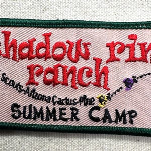 May include: Embroidered patch with the words "Shadow Rim Ranch" in red, arched over "Summer Camp" in black. The patch has a pink background, green border, and includes text for Girl Scouts Arizona Cactus Pine.
