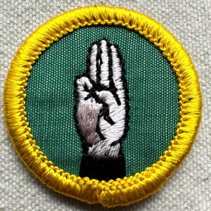 Pode incluir: Remendo bordado com um gesto de m&atilde;o com os dedos separados. A m&atilde;o &eacute; branca com contornos e detalhes pretos, sobre um fundo verde. O remendo redondo tem uma borda amarela.