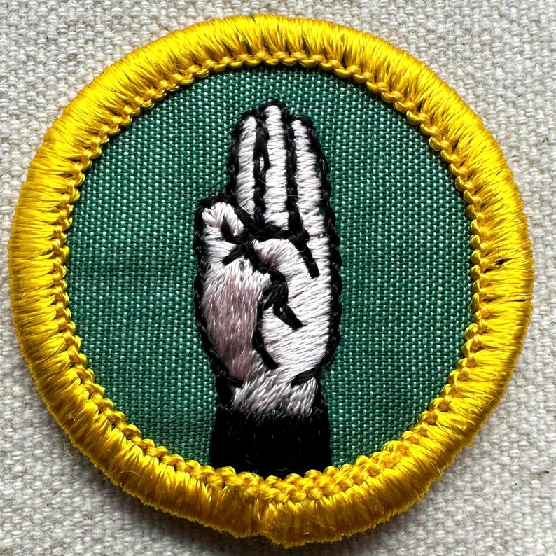Pode incluir: Remendo bordado com um gesto de m&atilde;o com os dedos separados. A m&atilde;o &eacute; branca com contornos e detalhes pretos, sobre um fundo verde. O remendo redondo tem uma borda amarela.
