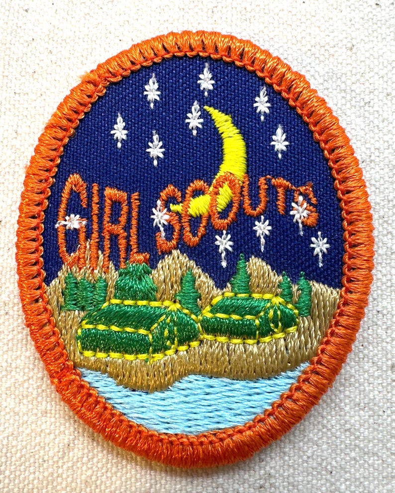 K&ouml;nnte beinhalten: Gestickter Girl Scouts-Aufn&auml;her mit orangefarbenem Rand. Der Aufn&auml;her zeigt einen blauen Nachthimmel mit wei&szlig;en Sternen und einem gelben Halbmond. Die Worte "GIRL SCOUTS" sind wei&szlig; gestickt. Darunter eine Landschaft mit gr&uuml;nen B&auml;umen und einem blauen Fluss.