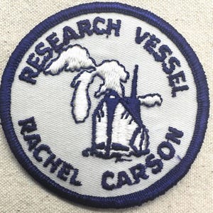 May include: A circular patch with a white background and a navy blue border. The patch features the words "RESEARCH VESSEL RACHEL CARSON" in navy blue, surrounding a stylized illustration of a ship and landmass in white and navy blue.