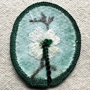 Op de afbeelding: Ovaalvormige geborduurde patch met een witte bloem met een groene stengel en accenten op een lichtblauwe achtergrond. De patch is omrand met donkergroene stiksels. De bloemblaadjes zijn gedetailleerd met witte draad en de stengel is een donkergroene vlecht.