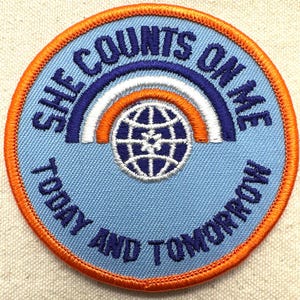 K&ouml;nnte beinhalten: Ein runder, gestickter Aufn&auml;her mit hellblauem Hintergrund und orangefarbenem Rand. Der Aufn&auml;her zeigt den Text "SHE COUNTS ON ME TODAY AND TOMORROW" in Dunkelblau. Ein Regenbogen-Design in Blau, Wei&szlig; und Orange w&ouml;lbt sich &uuml;ber einer Globus-Grafik.