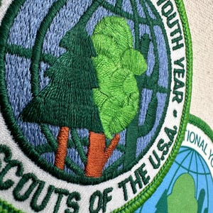 Op de afbeelding: Geborduurde patch met een groen boom- en wereldbolontwerp, met de tekst "SCOUTS OF THE U.S.A." en "INTERNATIONAL YOUTH YEAR." De patch heeft een witte achtergrond met groene en blauwe accenten.