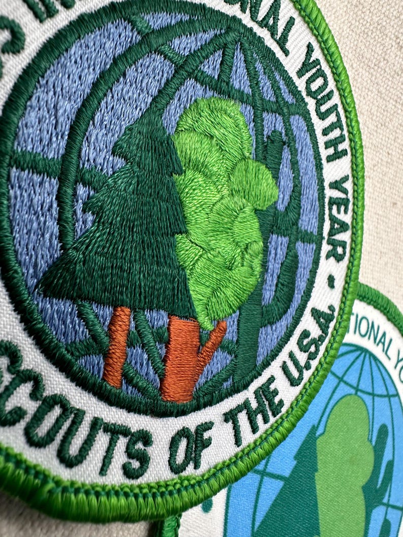 Op de afbeelding: Geborduurde patch met een groen boom- en wereldbolontwerp, met de tekst "SCOUTS OF THE U.S.A." en "INTERNATIONAL YOUTH YEAR." De patch heeft een witte achtergrond met groene en blauwe accenten.