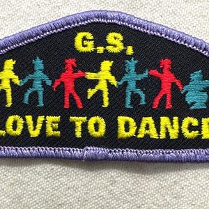 May include: Embroidered patch with a black background and a purple border. The patch features the text "G.S." and "LOVE TO DANCE" in yellow. Colorful figures of people holding hands are in the center. The figures are yellow, teal, red, and teal.