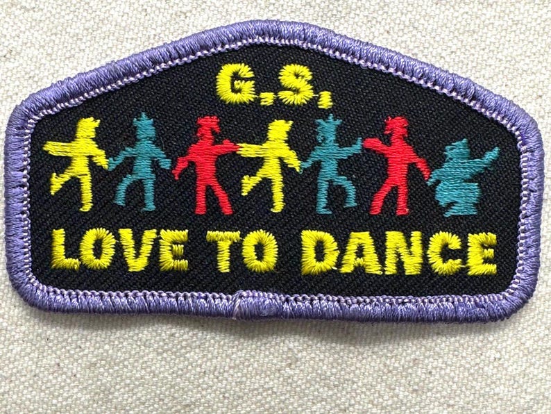 May include: Embroidered patch with a black background and a purple border. The patch features the text "G.S." and "LOVE TO DANCE" in yellow. Colorful figures of people holding hands are in the center. The figures are yellow, teal, red, and teal.