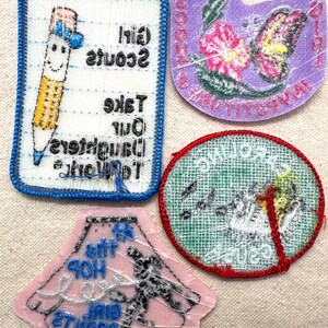 May include: Four Girl Scout patches with various designs. One patch features a smiling pencil with the text "Girl Scouts Take Our Daughters To The Top". Another shows a butterfly and flowers. A third patch has a girl dancing. The last patch has a bird.