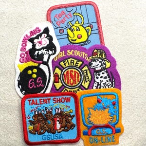 May include: A collection of colorful Girl Scout patches. Designs include a cat bowling, a teapot, a dalmatian dog firefighter, a talent show with bunnies, and an online theme. Each patch features embroidered details and text.
