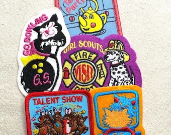 May include: A collection of colorful Girl Scout patches. Designs include a cat bowling, a teapot, a dalmatian dog firefighter, a talent show with bunnies, and an online theme. Each patch features embroidered details and text.