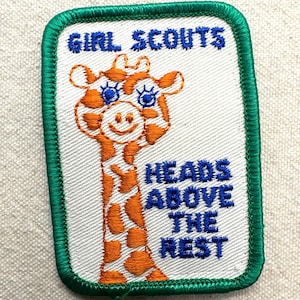 May include: Embroidered Girl Scouts patch with a giraffe design. The patch features the words "Girl Scouts" at the top and "Heads Above The Nest" below the giraffe. The giraffe is orange and white with blue accents, and the patch has a green border.