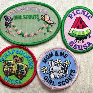 May include: Four Girl Scout patches with embroidered designs. One patch features butterflies and the words "Investiture Girl Scouts." Another shows a watermelon slice and the word "Picnic." A third depicts a bear reading a book, and the fourth shows bunnies and flowers.
