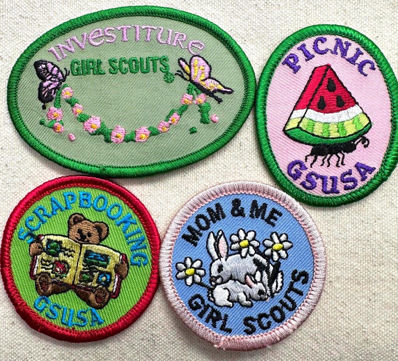 May include: Four Girl Scout patches with embroidered designs. One patch features butterflies and the words "Investiture Girl Scouts." Another shows a watermelon slice and the word "Picnic." A third depicts a bear reading a book, and the fourth shows bunnies and flowers.