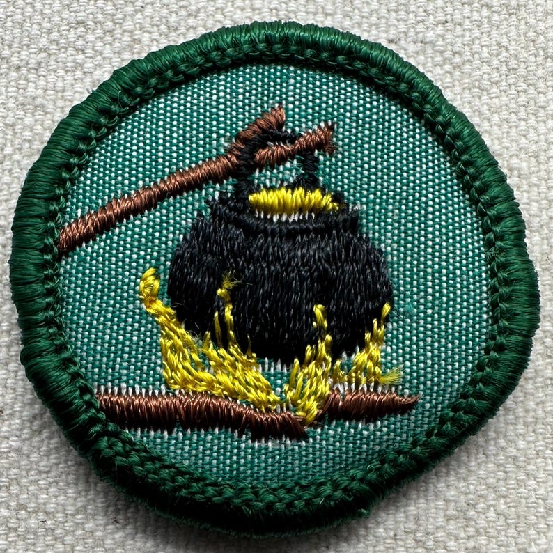 Puede incluir: Parche bordado con un caldero negro sobre un fuego amarillo y marr&oacute;n, con un palo marr&oacute;n encima. El parche tiene un fondo verde y un borde verde cosido. El parche es redondo.