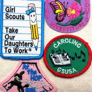 May include: A collection of Girl Scout patches with embroidered designs. One patch features a pencil with a bow and the text "Girl Scouts Take Our Daughters To Work." Other patches include a butterfly, musical notes, and a dancer.