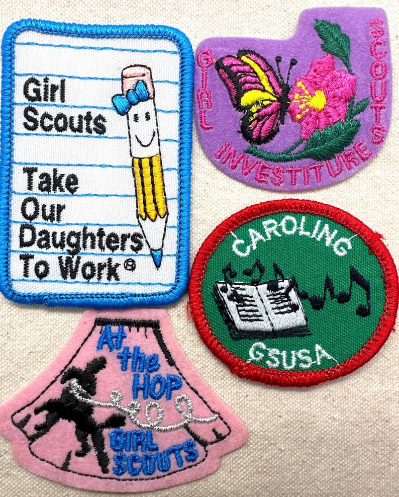 May include: A collection of Girl Scout patches with embroidered designs. One patch features a pencil with a bow and the text "Girl Scouts Take Our Daughters To Work." Other patches include a butterfly, musical notes, and a dancer.