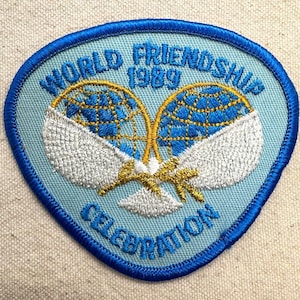 K&ouml;nnte beinhalten: Blau, wei&szlig; und gold bestickter Aufn&auml;her mit dem Text "WORLD FRIENDSHIP 1989 CELEBRATION". Der Aufn&auml;her zeigt zwei Tennisschl&auml;ger mit einem Globus-Design. Der Aufn&auml;her hat einen blauen Rand und einen hellblauen Hintergrund.