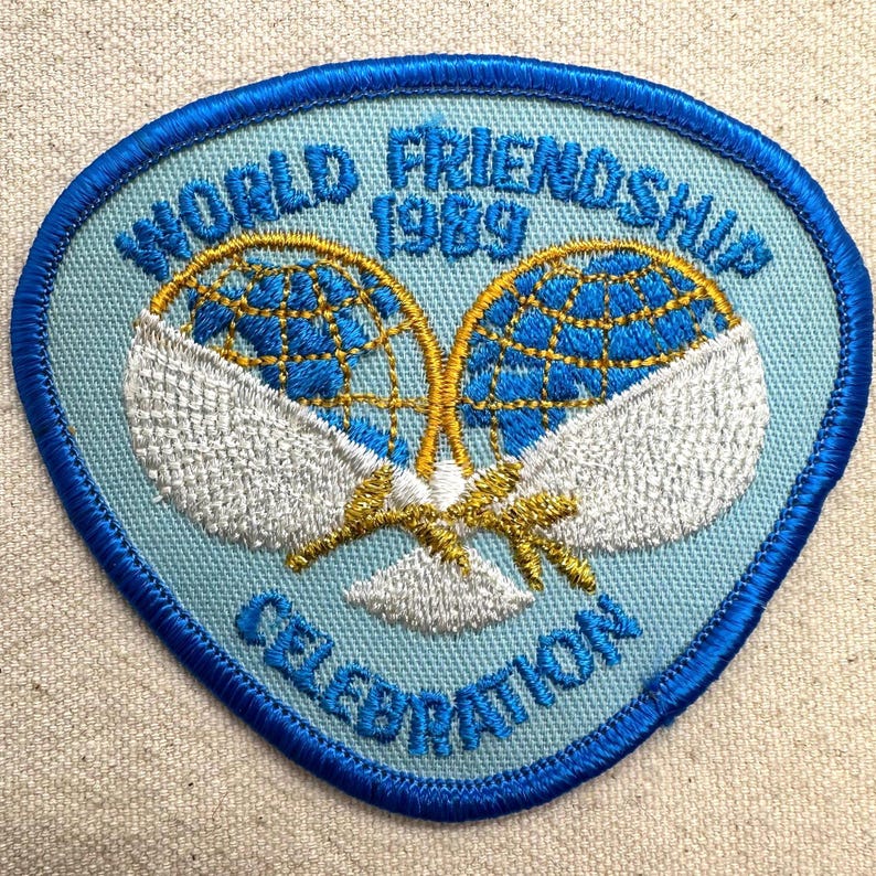 K&ouml;nnte beinhalten: Blau, wei&szlig; und gold bestickter Aufn&auml;her mit dem Text "WORLD FRIENDSHIP 1989 CELEBRATION". Der Aufn&auml;her zeigt zwei Tennisschl&auml;ger mit einem Globus-Design. Der Aufn&auml;her hat einen blauen Rand und einen hellblauen Hintergrund.