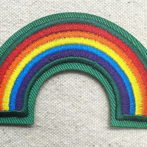 May include: Embroidered rainbow patch with a green border. The rainbow features bands of red, orange, yellow, blue, and purple. The patch is in the shape of an arch, and is likely for clothing or accessories.
