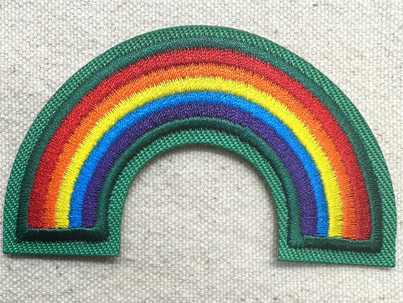 May include: Embroidered rainbow patch with a green border. The rainbow features bands of red, orange, yellow, blue, and purple. The patch is in the shape of an arch, and is likely for clothing or accessories.