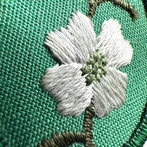 Op de afbeelding: Close-up van een geborduurd bloemmotief. Een witte bloem met een groen hart is op een groene stof gestikt. De bloem is omgeven door groene bladeren en stelen, allemaal geborduurd met gedetailleerd draadwerk. De achtergrond is een groene stof met textuur.