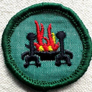 May include: A circular green embroidered patch featuring a campfire scene. The design includes a black fire pit with red, yellow, and orange flames, flanked by two black supports. The patch has a green border.