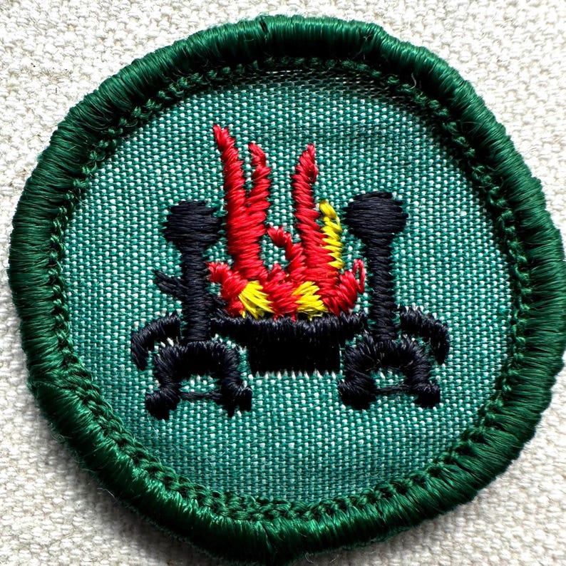 May include: A circular green embroidered patch featuring a campfire scene. The design includes a black fire pit with red, yellow, and orange flames, flanked by two black supports. The patch has a green border.