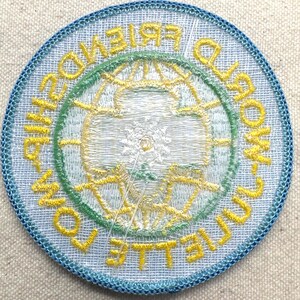 Peut inclure: &Eacute;cusson brod&eacute; circulaire avec une bordure bleu clair. Le motif central repr&eacute;sente un globe et les mots "WORLD FRIENDSHIP TOWN COLLEGIATE" en jaune et vert. L'&eacute;cusson a un aspect textur&eacute;.