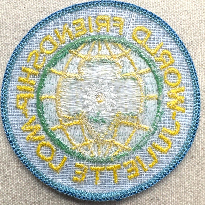 Peut inclure: &Eacute;cusson brod&eacute; circulaire avec une bordure bleu clair. Le motif central repr&eacute;sente un globe et les mots "WORLD FRIENDSHIP TOWN COLLEGIATE" en jaune et vert. L'&eacute;cusson a un aspect textur&eacute;.