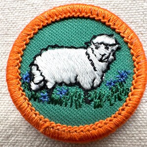May include: Embroidered patch featuring a white sheep on a green background, surrounded by orange stitching. The sheep is depicted in a detailed, textured style, with blue flowers and green grass at its feet. A charming, pastoral design.